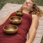 Woman lying on a mat with three bronze singing bowls in front of her, with prayer flags in the background.