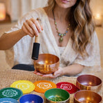 Woman holding a copper singing bowl with various colored bowls and their corresponding chakras on a table.