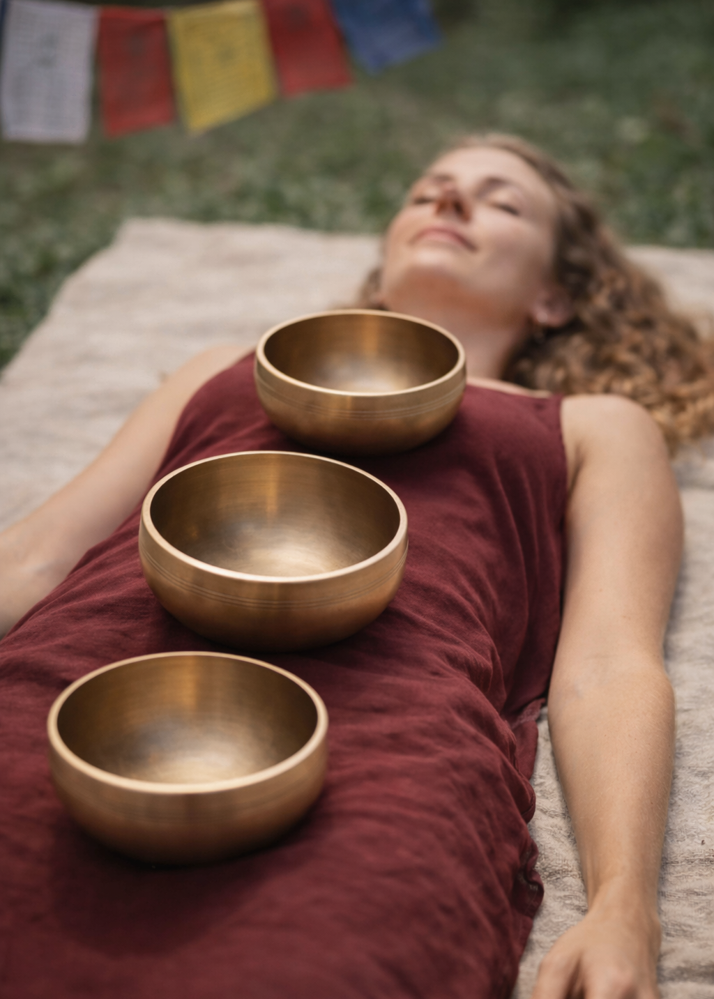 Woman lying on a mat with three bronze singing bowls in front of her, with prayer flags in the background.