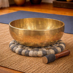 Brass singing bowl with a wooden handle on a textured mat in a room setting.