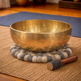 Brass singing bowl with a wooden handle on a textured mat in a room setting.