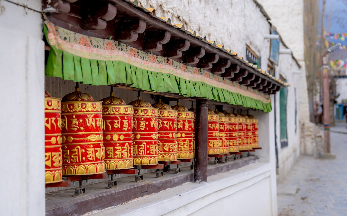 A row of traditional Tibetan prayer wheels hanging under a green fringed awning on a white building wall, with a blurred street scene in the background.