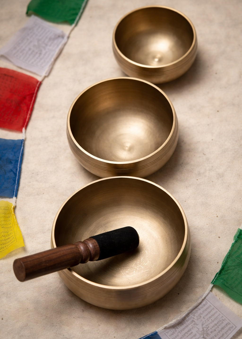 Three brass singing bowls on a textured surface with prayer flags in the background