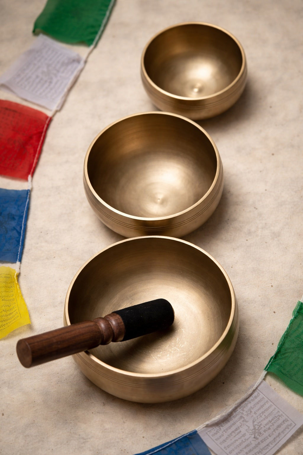 Three brass singing bowls on a textured surface with prayer flags in the background
